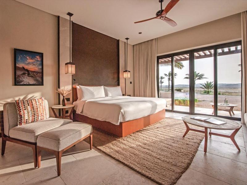 Chambre d'hôtel moderne avec grand lit, sièges et vue sur la mer à travers des fenêtres.