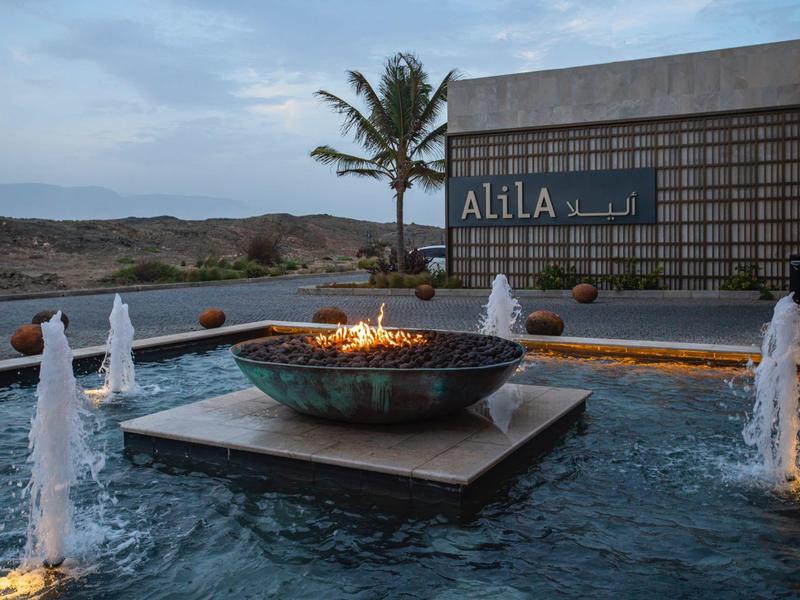 Espace piscine moderne avec brasero et fontaines devant un mur d'hôtel au crépuscule.