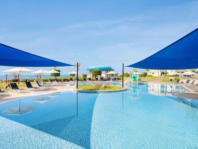 Groot zwembad met blauwe zonneschermen, ligstoelen en uitzicht op zee bij heldere hemel.