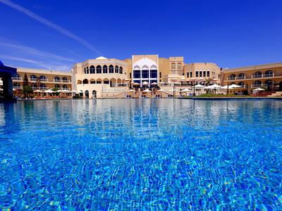 Großer blauer Hotelpool mit hellen Gebäuden und klarem Himmel im Hintergrund.