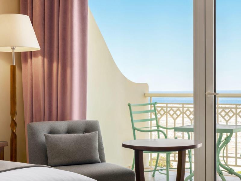 Lichte hotelkamer met balkon, stoel, tafel, fauteuil en uitzicht op de blauwe lucht.