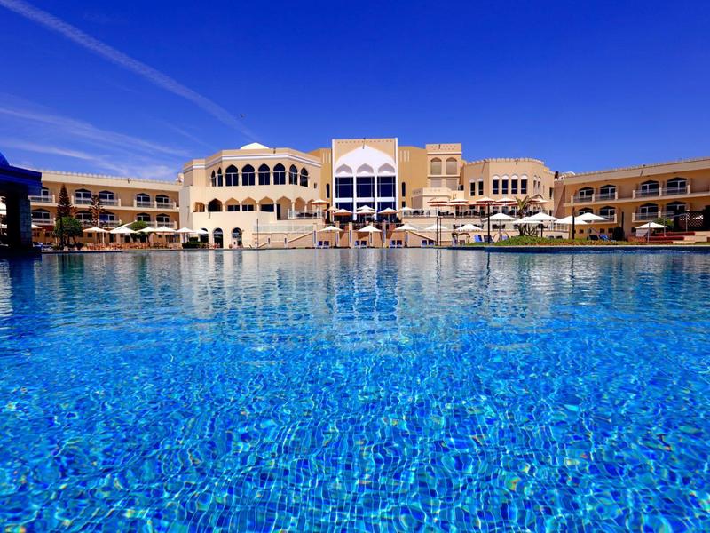 Groot zwembad met helder blauw water voor een luxe hotel onder een blauwe lucht.