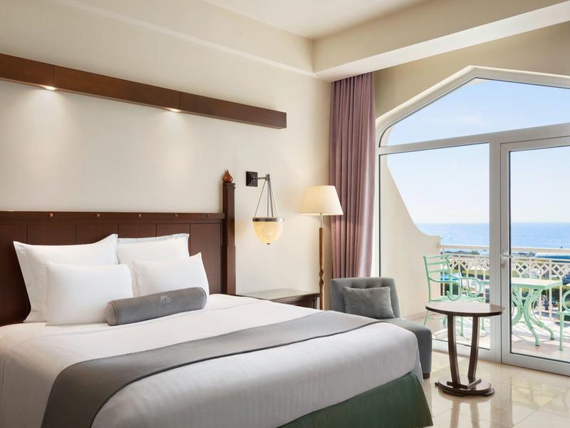 Moderne hotelkamer met groot bed, zithoek en zeezicht door ramen van vloer tot plafond.