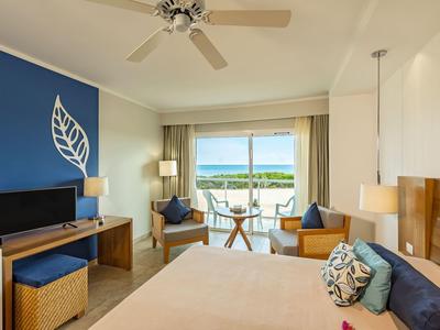 Hell eingerichtetes Hotelzimmer mit Balkon, Doppelbett, Fernseher, Stühlen und Blick auf Wasser.
