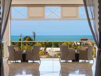 Balkon mit Stühlen und Tischen, Blick auf Pflanzen, Palmen und dahinter das blaue Meer.