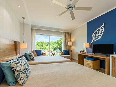 Helles Hotelzimmer mit zwei Betten, blauem Akzent-Wand, Deckenventilator und Blick auf Grünflächen.