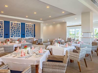 Helles Restaurant mit gedeckten Tischen, weißen Tischdecken und gepolsterten Stühlen aus Rattan.