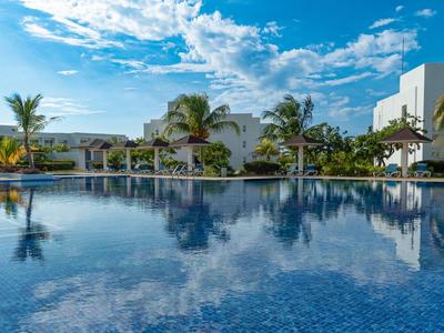 Großer blauer Pool mit Spiegelung des Himmels, umgeben von Palmen und weißen Gebäuden.