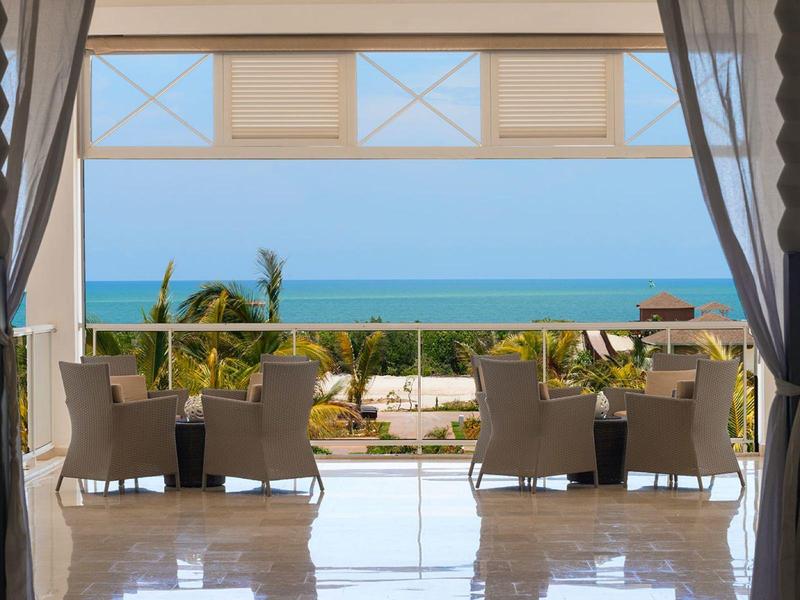 Balkon mit Stühlen und Tischen, Blick auf Pflanzen, Palmen und dahinter das blaue Meer.