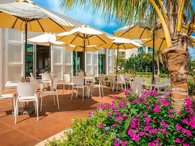 Außenbereich eines Cafés mit weißen Stühlen, großen Sonnenschirmen und bunter Blumenrabatte.