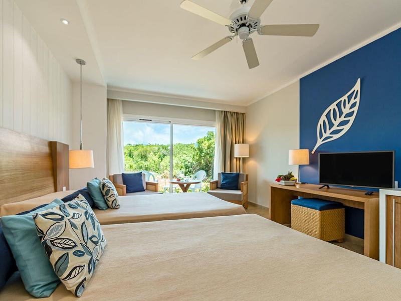 Helles, modernes Hotelzimmer mit blauen Akzenten, zwei Betten, TV und großer Glasfront zum Garten.