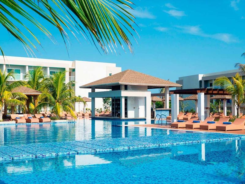 Ein großes Schwimmbecken mit blauen Fliesen, umgeben von Liegestühlen, Palmen und modernem Gebäude.