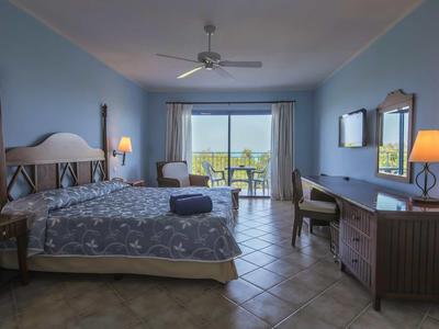 Hellblaues Hotelzimmer mit großem Bett, Schreibtisch, Stuhl, Balkon und Deckenventilator.