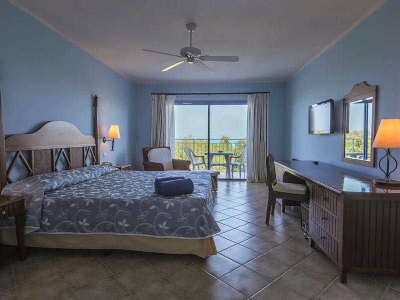 Hellblaues Hotelzimmer mit großem Bett, Schreibtisch, Stuhl, Balkon und Deckenventilator.