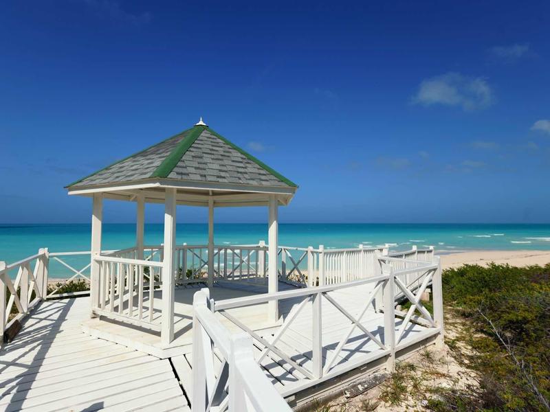Weißes Holzdeck mit Pavillon am Strand, türkisfarbenes Meer und klarer blauer Himmel im Hintergrund.