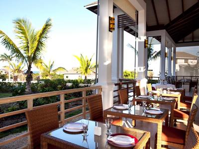 Offenes Restaurant mit Holztischen, Stühlen, Palmen und Blick auf einen Garten bei Sonnenschein.