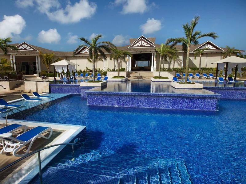 Großer blauer Pool mit Liegestühlen und Sonnenschirmen vor einem Gebäudekomplex bei sonnigem Himmel.