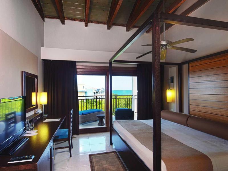 Hotelzimmer mit Doppelbett, Holzmöbeln, TV, Schreibtisch und Balkon mit Meerblick.