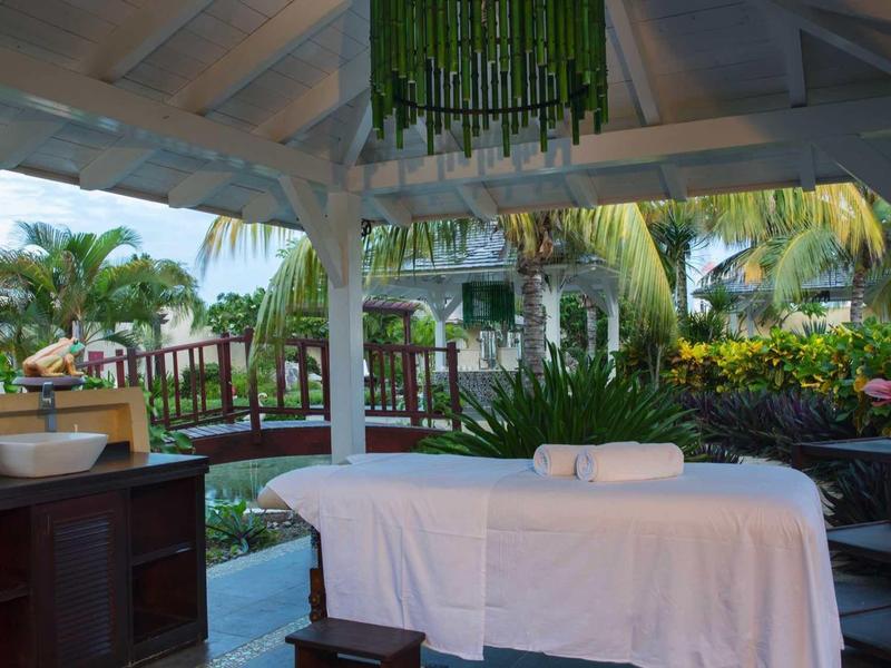 Offener Massagebereich mit weißer Liege, grünen Pflanzen und tropischem Ambiente unter Holzdecke.