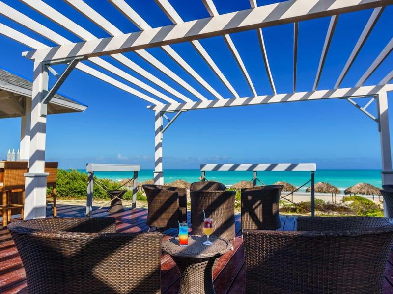 Terrasse mit Rattanmöbeln, blau-weißer Markise und Blick aufs Meer unter strahlend blauem Himmel.