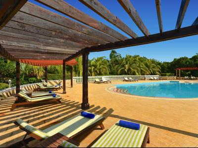 Poolbereich mit Liegestühlen unter Holzdach, blauem Wasser und grünen Palmen im Hintergrund.