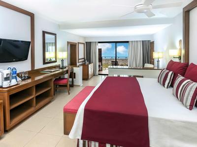 Hell eingerichtetes Hotelzimmer mit großem Bett, rotem Bettläufer, TV und Blick aufs Meer.