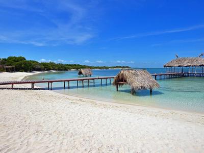 Weißer Sandstrand mit Steg und Hütten über klarem, türkisfarbenem Wasser.