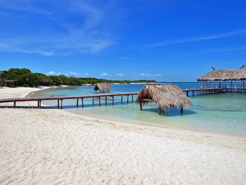Weißer Sandstrand, blauer Himmel, Holzsteg und kleine Hütten über klarem Meerwasser.