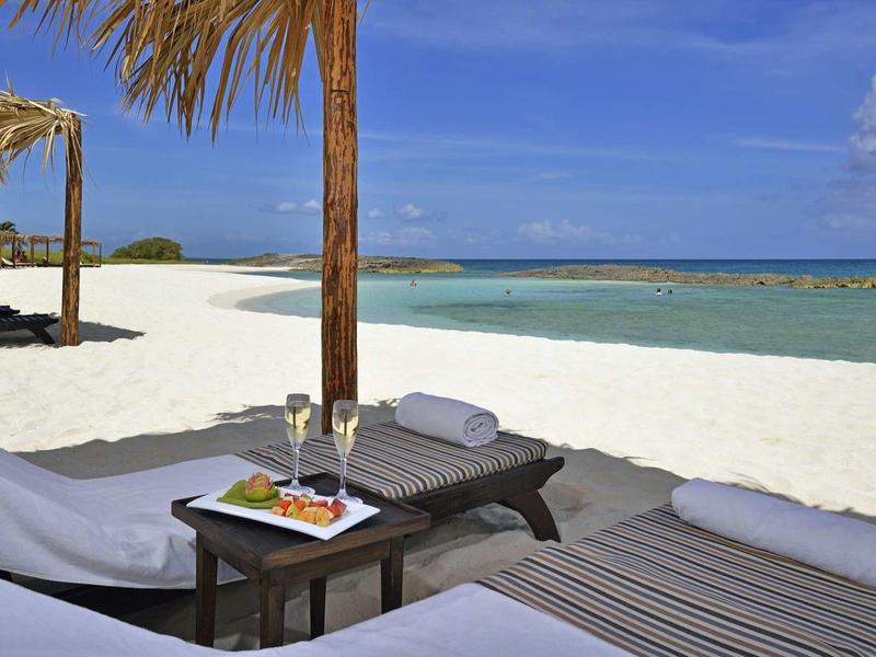 Strand mit weißen Liegen, Holztisch, Snacks und türkisblauem Meer unter blauem Himmel.