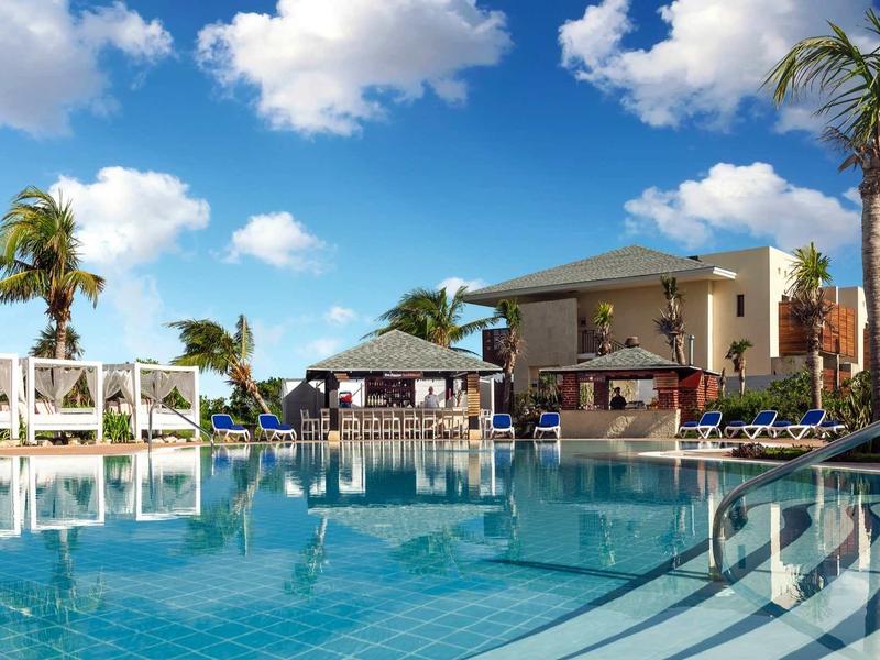 Großer blauer Pool mit Liegen, Palmen und Gebäuden unter blauem Himmel mit weißen Wolken.