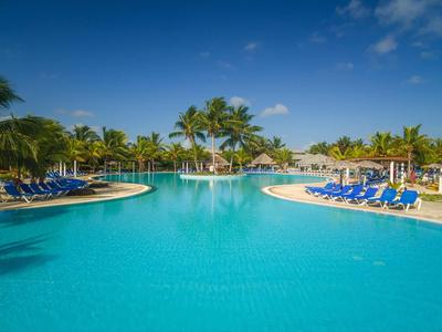 Großer Pool mit klar blauem Wasser, umgeben von Palmen und Sonnenschirmen mit Liegestühlen.