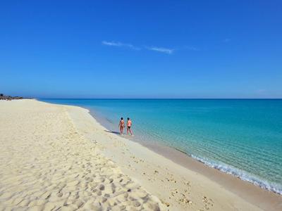 Strand mit hellem Sand, blauem Himmel und türkisfarbenem Meer, zwei Personen am Wasser.