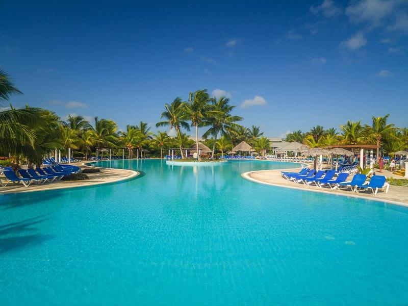 Großer Pool mit klarem blauem Wasser, umgeben von Palmen und blauen Liegestühlen unter Sonnenschirmen.