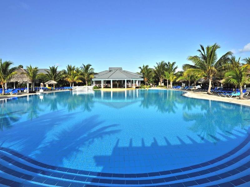 Großer blauer Swimmingpool mit Palmen und Sonnenschirmen unter klarem Himmel.