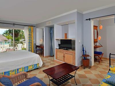 Helles Wohnzimmer mit Sofa, Couchtisch, Fernseher, angrenzendem Schlafzimmer und Balkon mit grünem Ausblick.