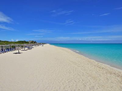 Breiter Sandstrand mit Liegestühlen, klarem türkisblauem Wasser und blauem Himmel mit wenigen Wolken.