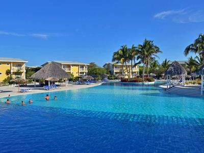 Großer Pool mit Palmen und gelben Gebäuden unter blauem Himmel in einem tropischen Resort.