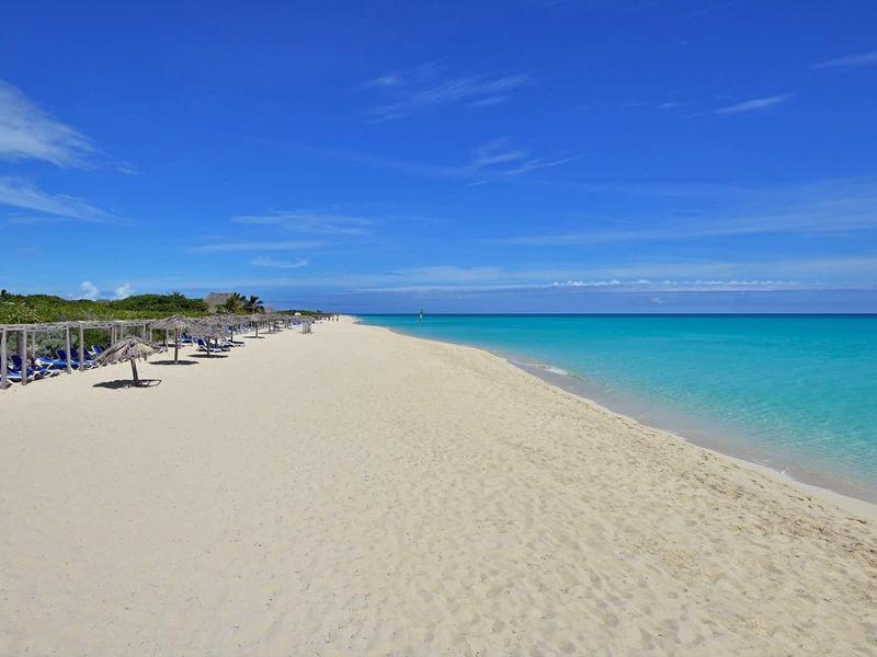 Breiter Sandstrand mit Liegestühlen, klarem türkisblauem Wasser und blauem Himmel mit wenigen Wolken.