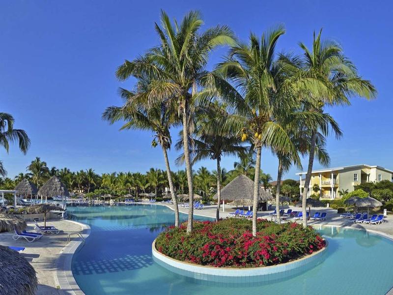 Großer Pool mit Palmeninsel, blauer Wasserfläche und Sonnenschirmen in sonnigem Resort.