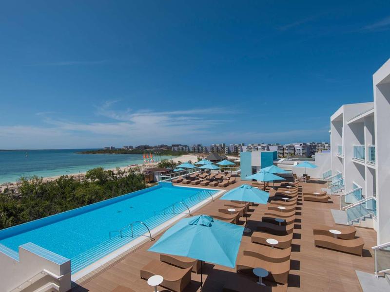 Moderner Poolbereich am Meer mit blauen Liegen, Schirmen und weißem Hotelgebäude bei klarem Himmel.