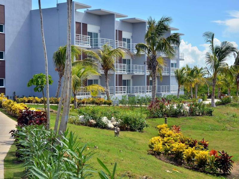 Modernes Hotelgebäude mit Balkonen, umgeben von grünen Palmen und bunten Blumenbeeten.