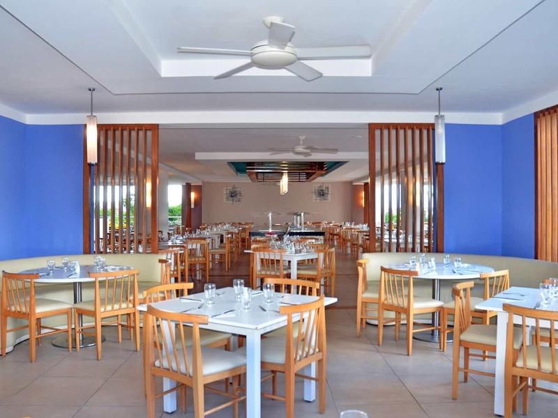 Hell eingerichteter Speisesaal mit Holzstühlen, Tischen und blauen Wänden. Deckenventilator an der Decke.