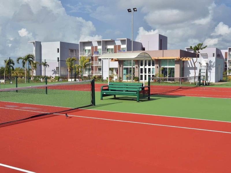 Roter Tennisplatz mit grünem Netz, Bank und Gebäuden im Hintergrund unter bewölktem Himmel.