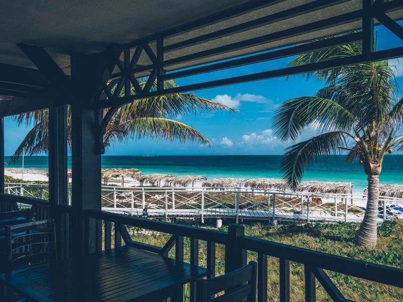 Blick von einer überdachten Terrasse mit Holzgeländer auf Palmen, Strand und blaues Meer unter blauem Himmel.