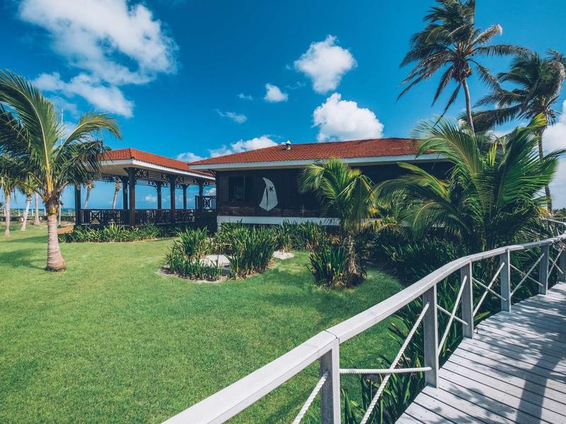 Bungalow mit rotem Dach, Palmen und grünem Rasen unter blauem Himmel mit weißen Wolken.