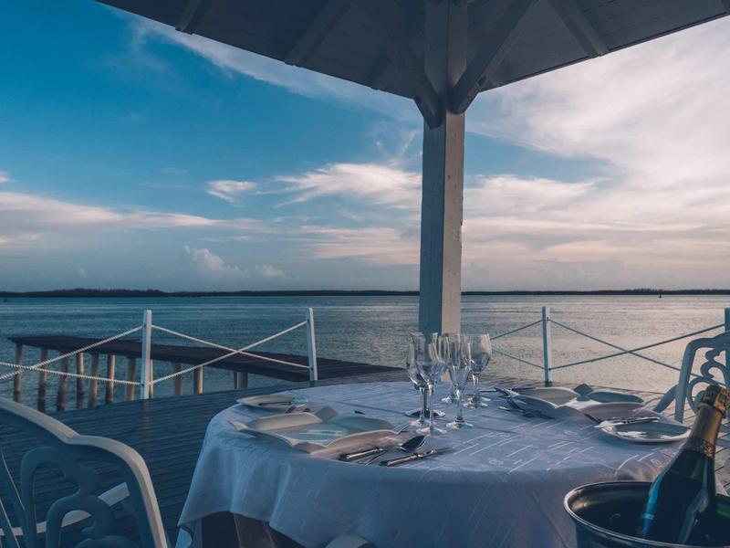 Gedeckter Tisch mit Gläsern und Besteck unter einem Pavillon am Wasser bei Sonnenuntergang.