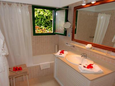 Helles Badezimmer mit weißen Fliesen, großem Holzbalkenspiegel und offenem Fenster, dekoriert mit roten Blumen.