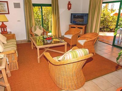Helles Wohnzimmer mit Rattanstühlen, orangem Teppich und großen Fenstern zum Garten.