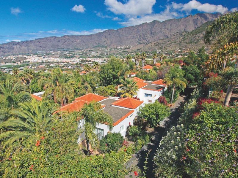 Panoramic view of green palm trees, houses with red roofs, and mountains under a blue sky.