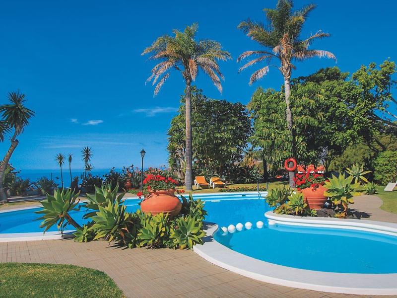 Pool area with palm trees and blooming plants under a clear blue sky.